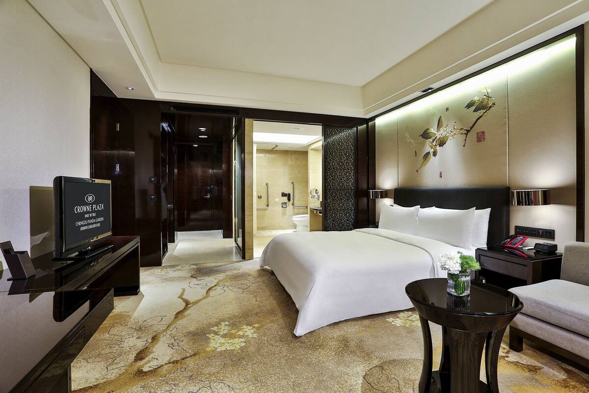 A hotel room with a large bed, a TV, a bedside table, a sofa, and a bathroom visible through an open door.