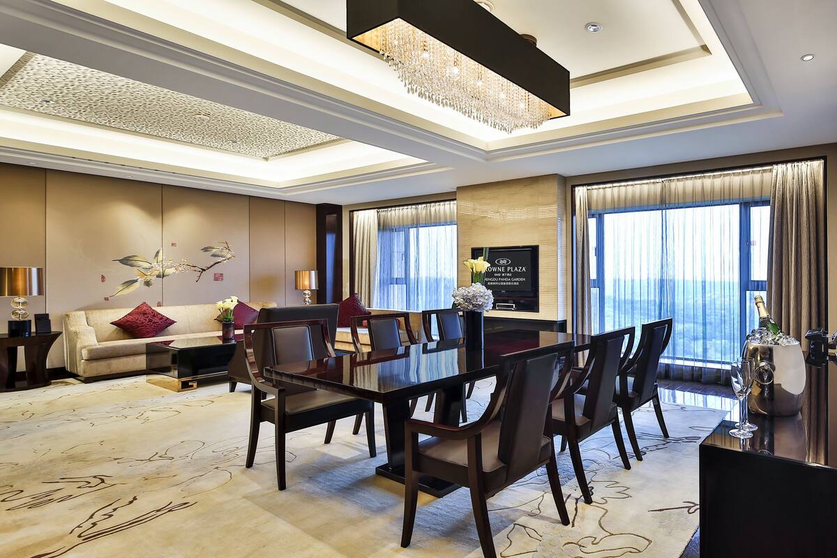 A spacious hotel room with a large dining table, comfortable seating, a sofa, and a large window offering a view.