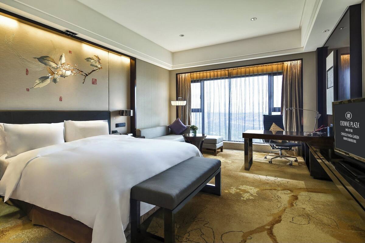 A hotel room with a large bed, a desk, a chair, a bench, a TV, and a large window with curtains.