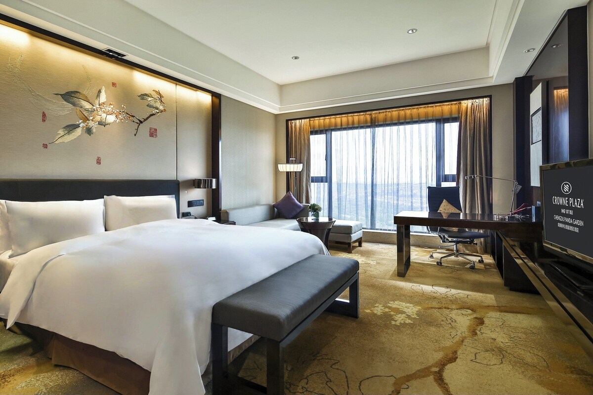 A hotel room with a large bed, a desk, a chair, a bench, a TV, and a large window with curtains.