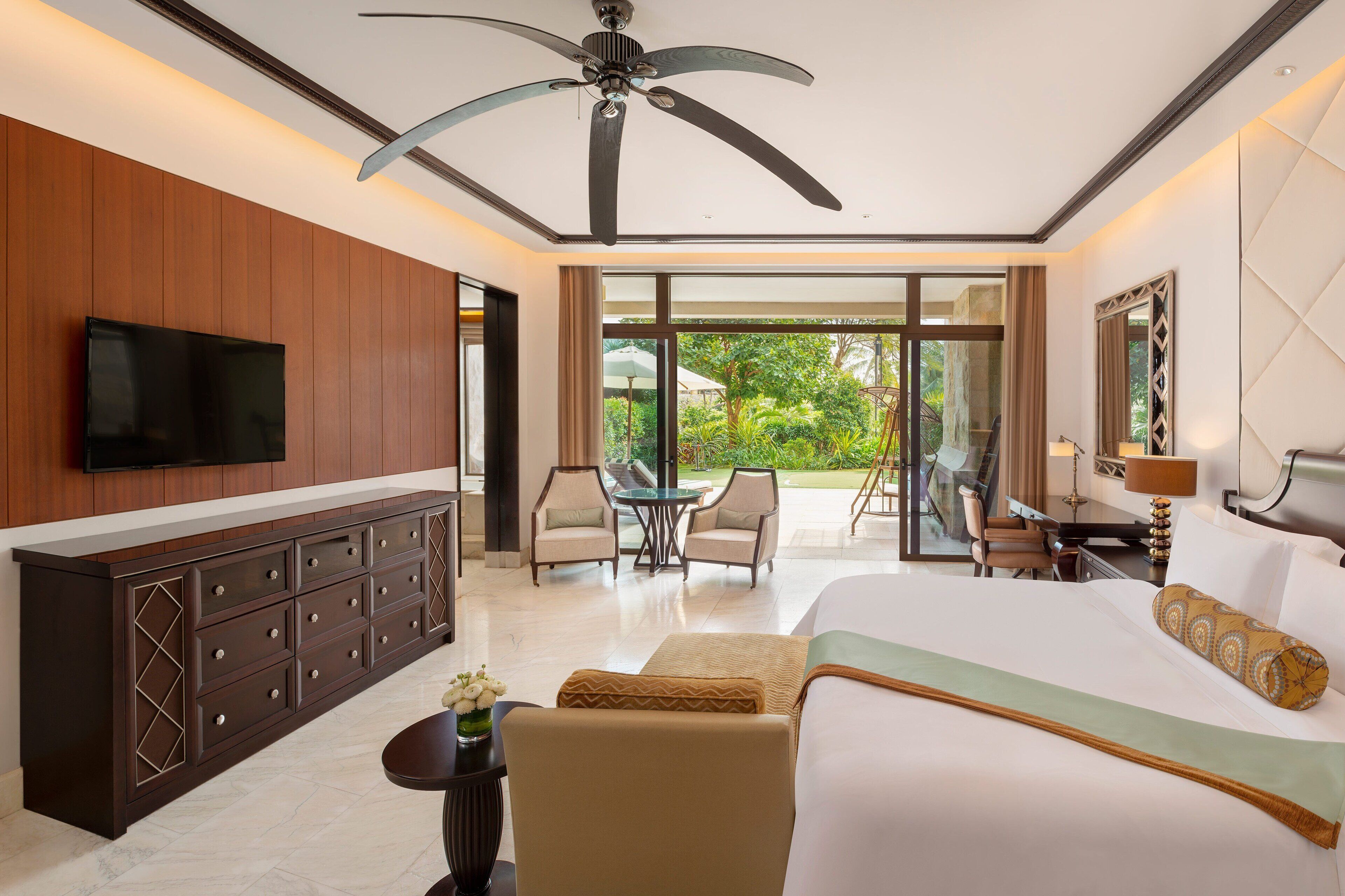 A hotel room with a large bed, a TV mounted on the wall, a ceiling fan, a dining area with chairs, and a view of the outdoors.