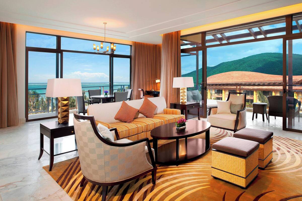 Une chambre d’hôtel spacieuse dotée d’une grande fenêtre offrant une vue sur la mer et les collines, d’un canapé confortable, d’une table à manger et d’un petit coin cuisine.