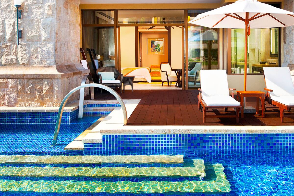 Un espace piscine avec des chaises longues et une table, un escalier menant à un espace de détente abrité et un mur en pierre.