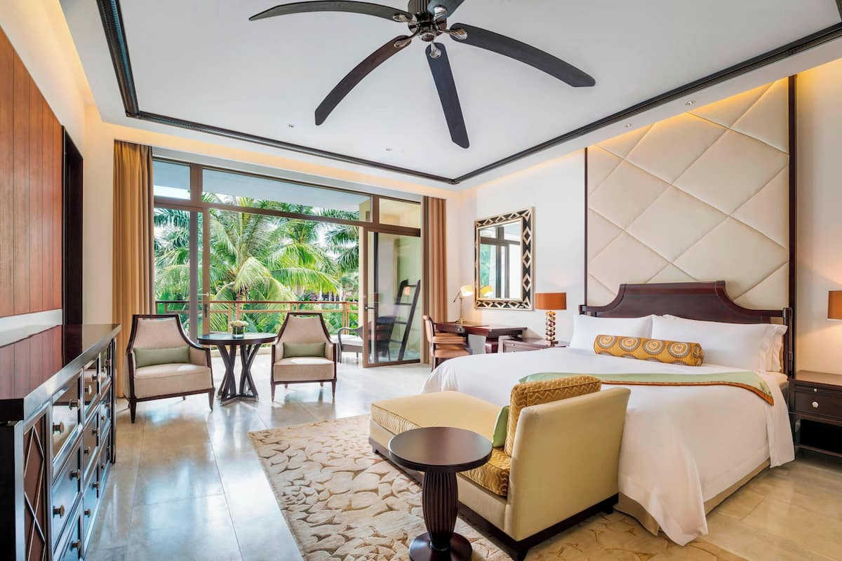 Une chambre d’hôtel équipée d’un lit, d’un bureau, d’une chaise, d’un canapé, d’un ventilateur de plafond et d’une grande fenêtre donnant sur des palmiers.