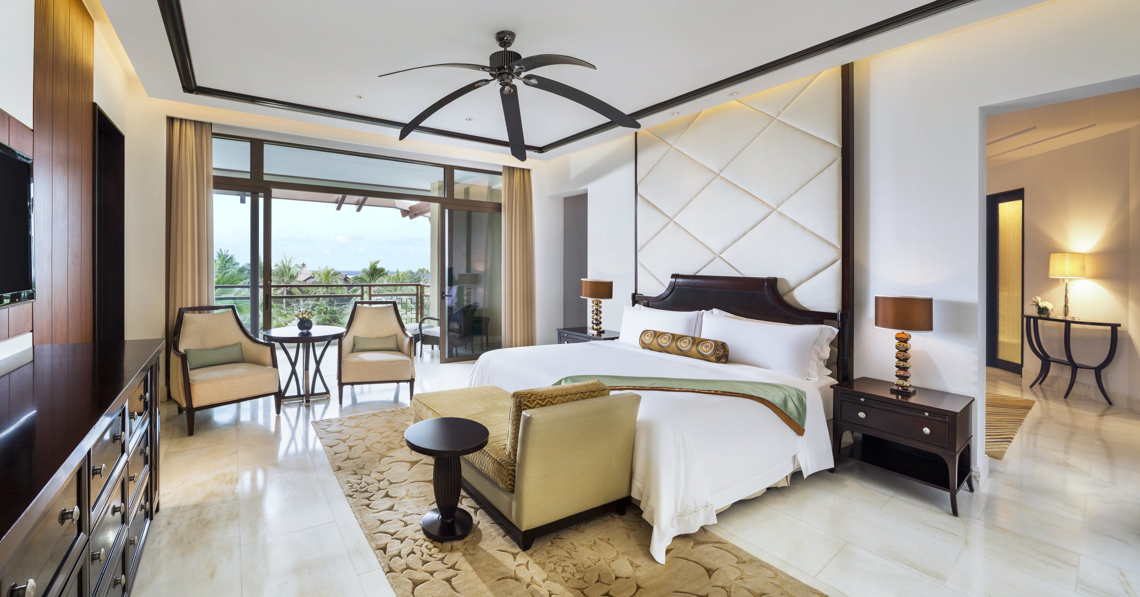 A modern hotel room with a large bed, a sitting area with a sofa and armchairs, a TV, and a balcony with a view.
