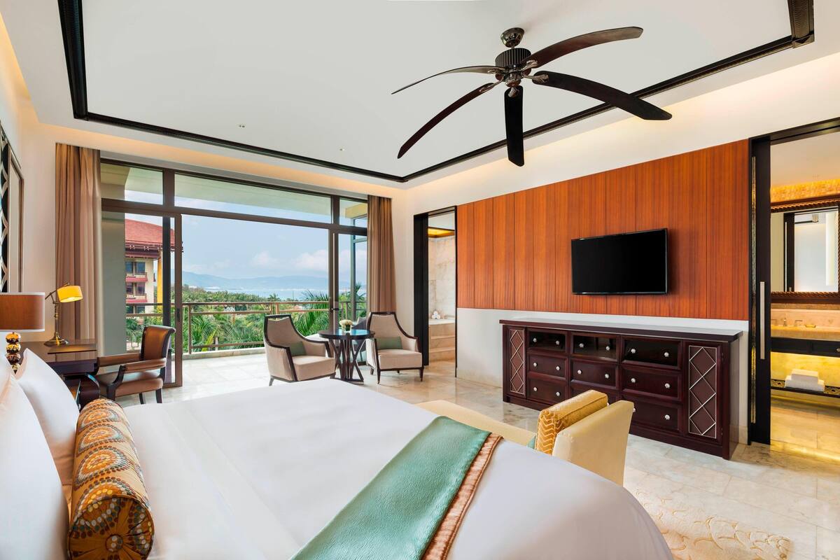 Une chambre d’hôtel dotée d’un grand lit, d’un ventilateur de plafond, d’un téléviseur fixé au mur, d’un mur recouvert de panneaux en bois, d’un balcon avec vue et d’un bureau avec une lampe.