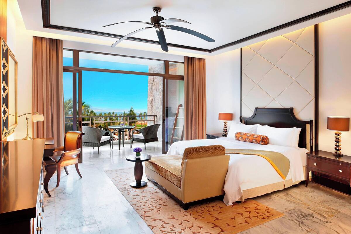 Une chambre d’hôtel moderne dotée d’un grand lit, d’un ventilateur de plafond, d’un balcon avec vue et d’un coin repas avec des chaises et une table.