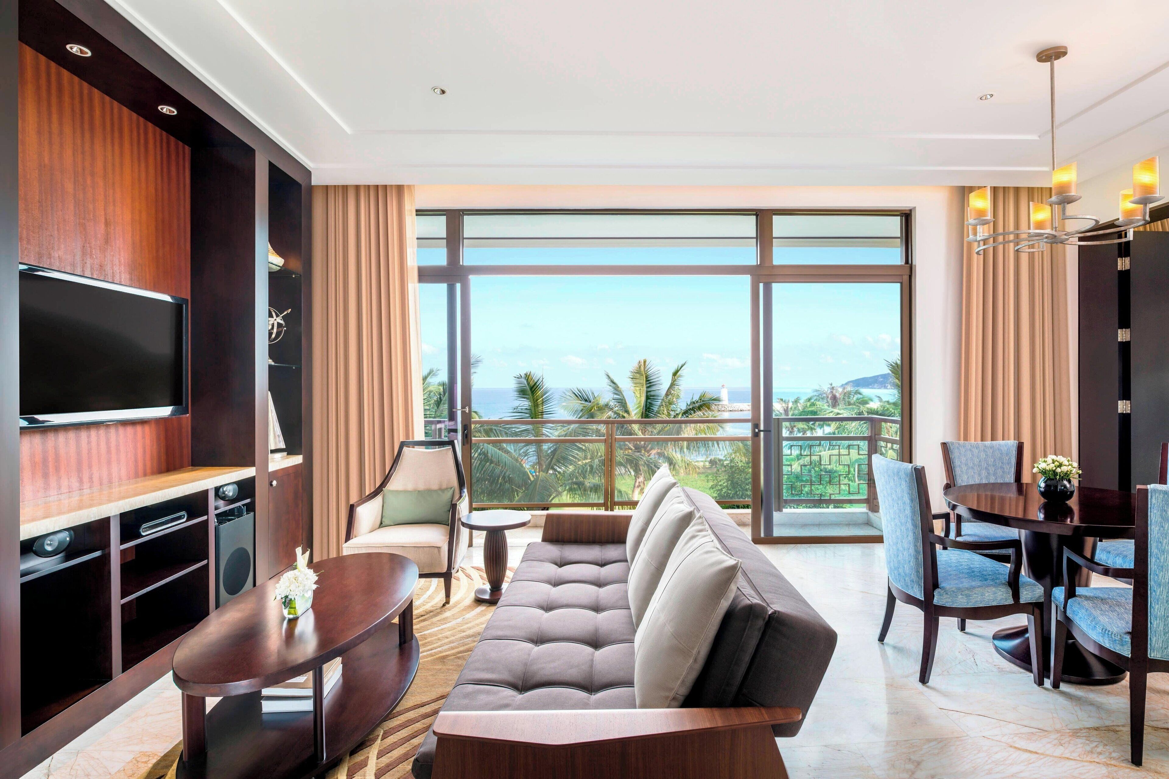 A modern living room with a large flat-screen TV, a sofa, a dining table with chairs, and a balcony with a view of the ocean.