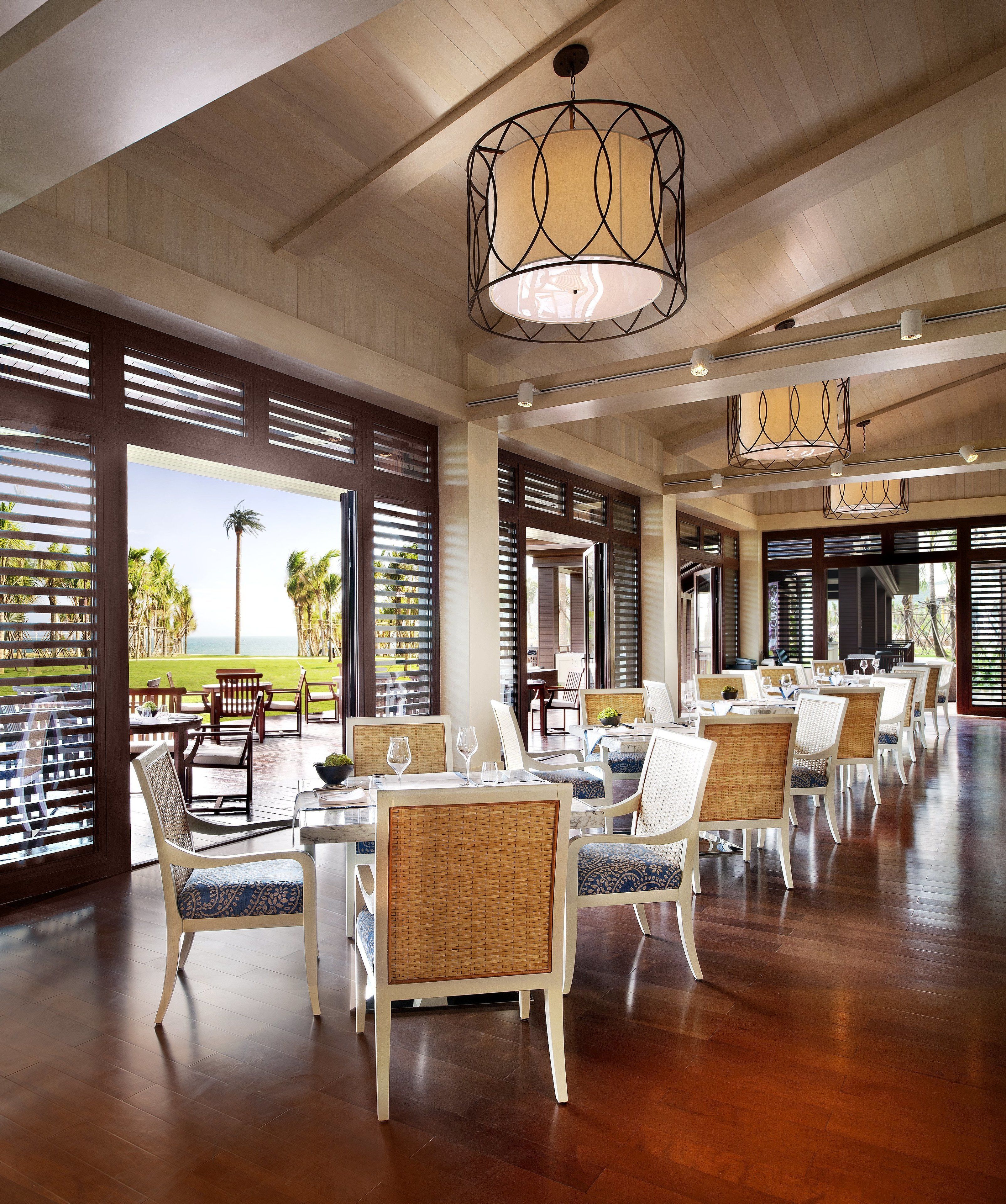 A spacious indoor dining area with wooden flooring and wicker chairs, large windows with blinds, and hanging pendant lights.