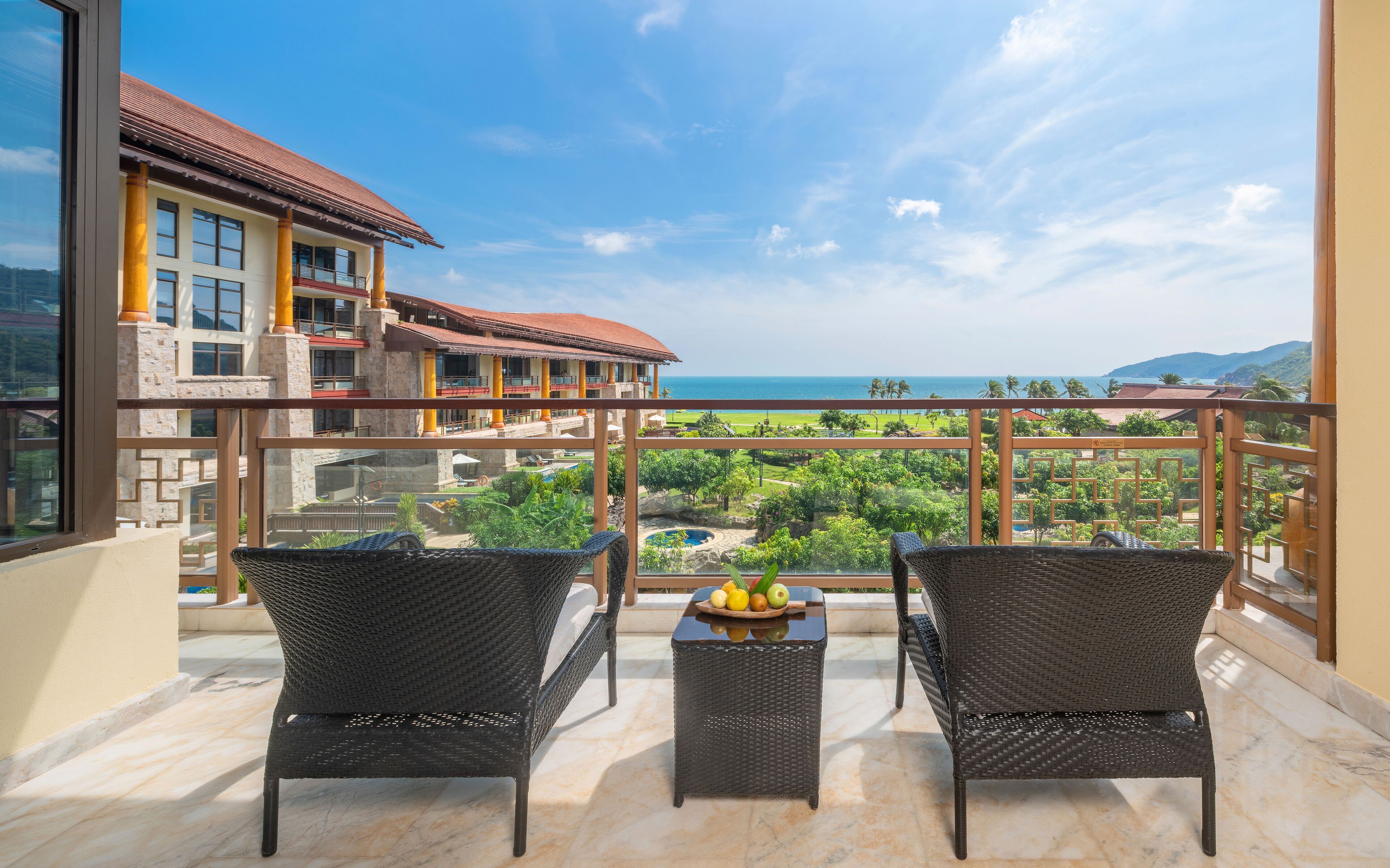 Ein Balkon mit Korbstühlen und einem Tisch, der auf einen Pool und üppiges Grün blickt, mit Blick auf das Meer und einem Hotelgebäude im Hintergrund.
