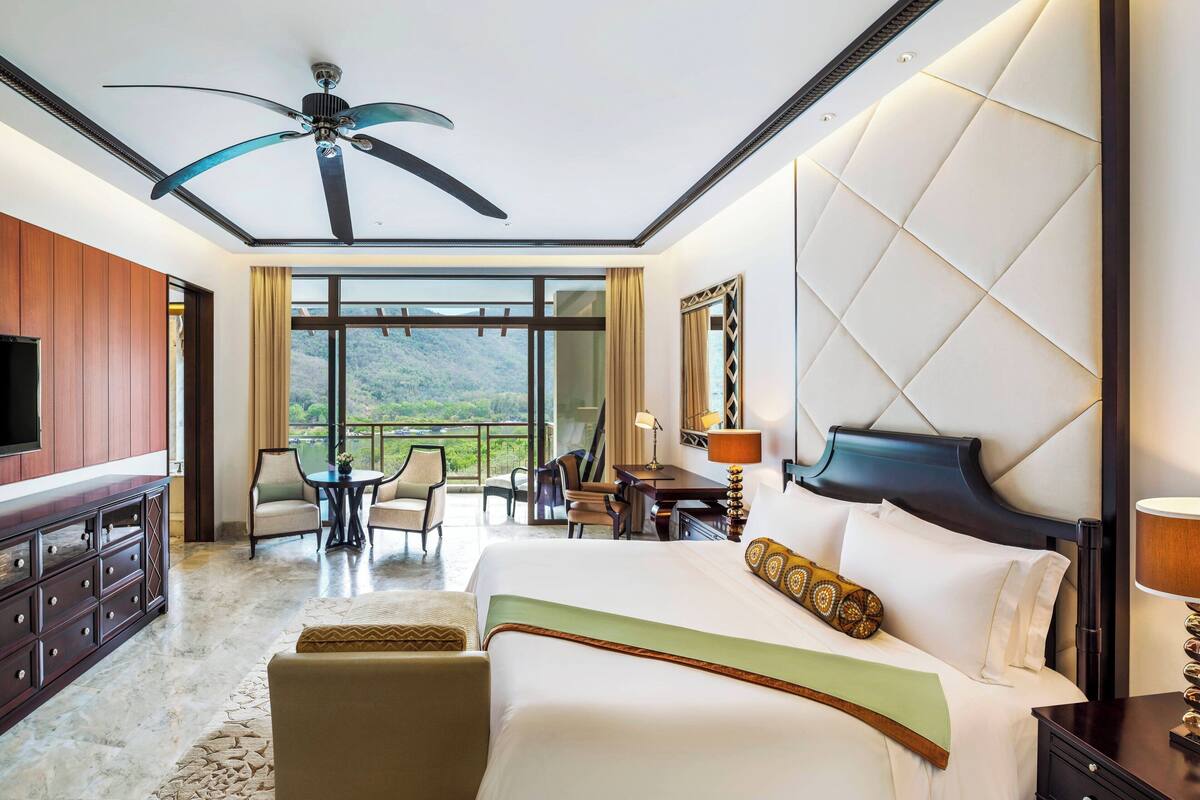 Une chambre d’hôtel dotée d’un grand lit, d’un ventilateur de plafond, d’une télévision, d’un balcon offrant une vue sur les montagnes, ainsi que d’un coin repas avec des chaises et une table.
