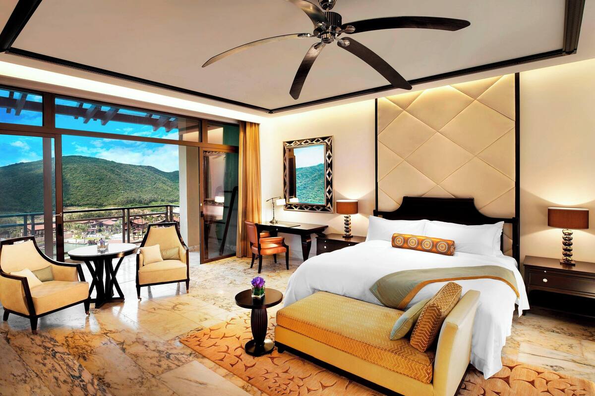 Une chambre d’hôtel comprenant un lit, deux fauteuils, une petite table avec un vase, un bureau et une chaise. Une grande fenêtre offre une vue sur les montagnes.