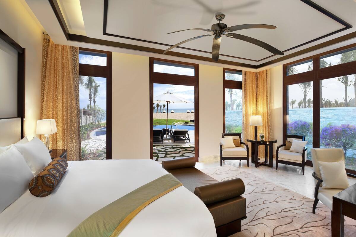 Une chambre d’hôtel avec un grand lit, un ventilateur de plafond et une vue sur une piscine et une plage.