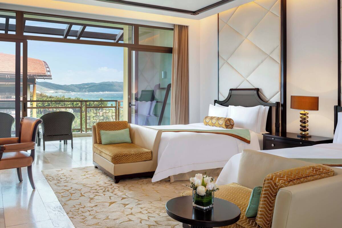 Une chambre d’hôtel avec deux lits, un canapé, un fauteuil, un bureau et un balcon donnant sur la mer.