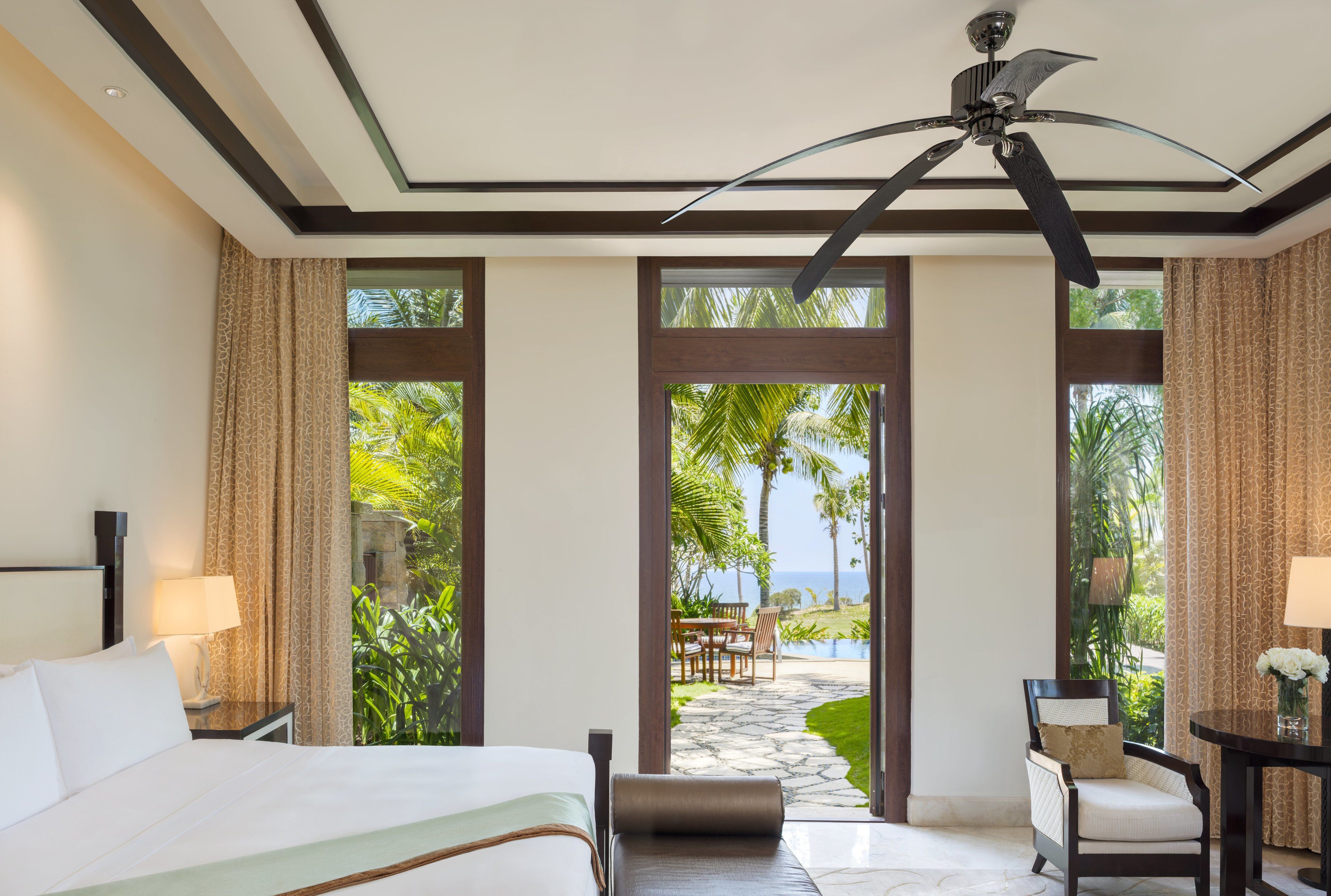 Ein Hotelzimmer mit Bett, Sessel, Tisch und Blick auf einen tropischen Garten und das Meer.