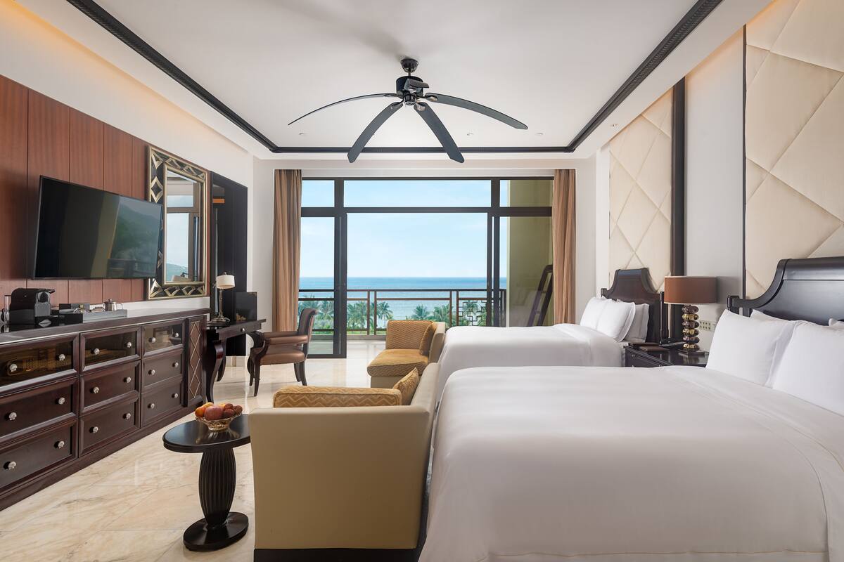 Une chambre d’hôtel dotée d’un grand lit, d’un bureau avec un téléviseur à écran plat, d’un coin repas avec une table et des chaises, et d’un balcon offrant une vue sur l’océan.
