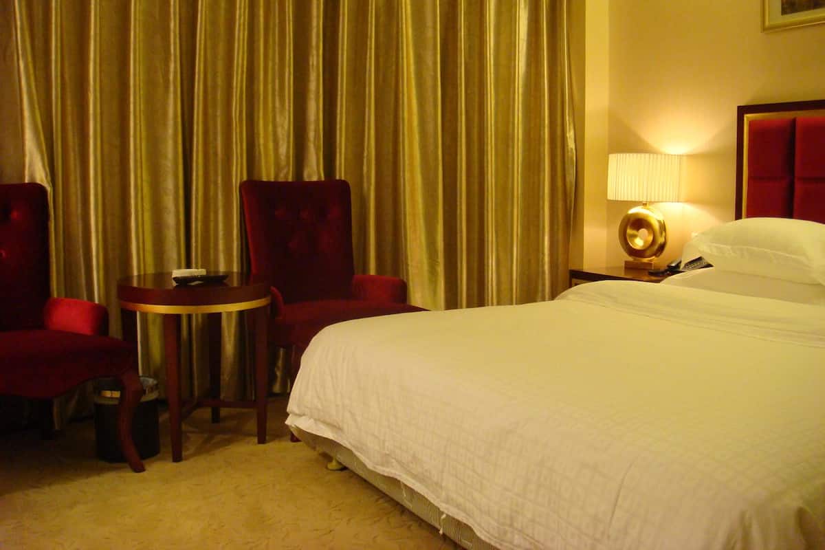 Een hotelkamer met een bed, twee rode stoelen, een tafeltje, een lamp en gordijnen.