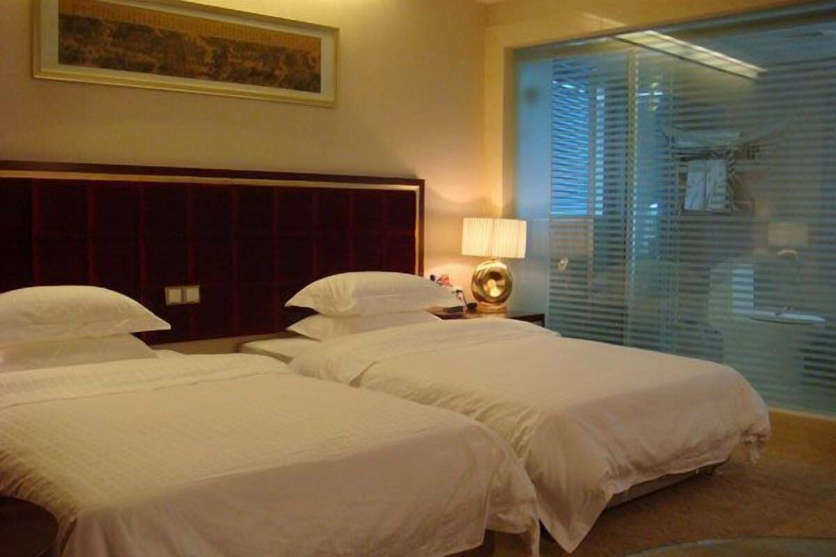 Hotelkamer met twee bedden, een hoofdeinde, een nachtkastje met een lamp en een groot raam met jaloezieën.