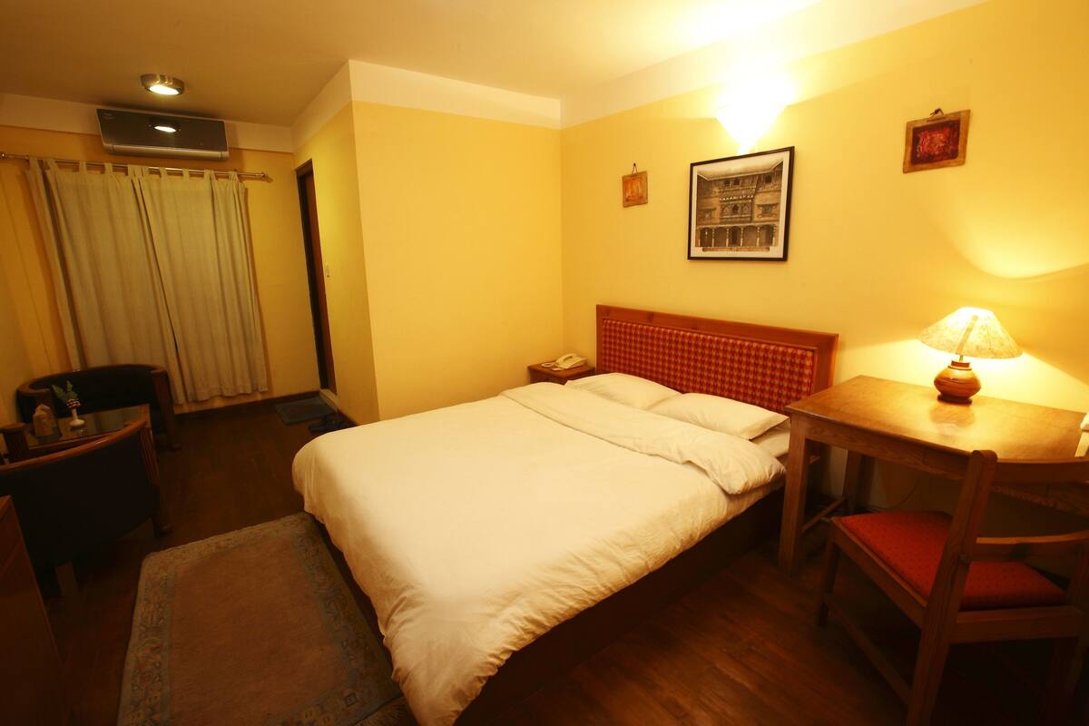 A hotel room with a bed, bedside table, lamp, chair, desk, and a picture on the wall.
