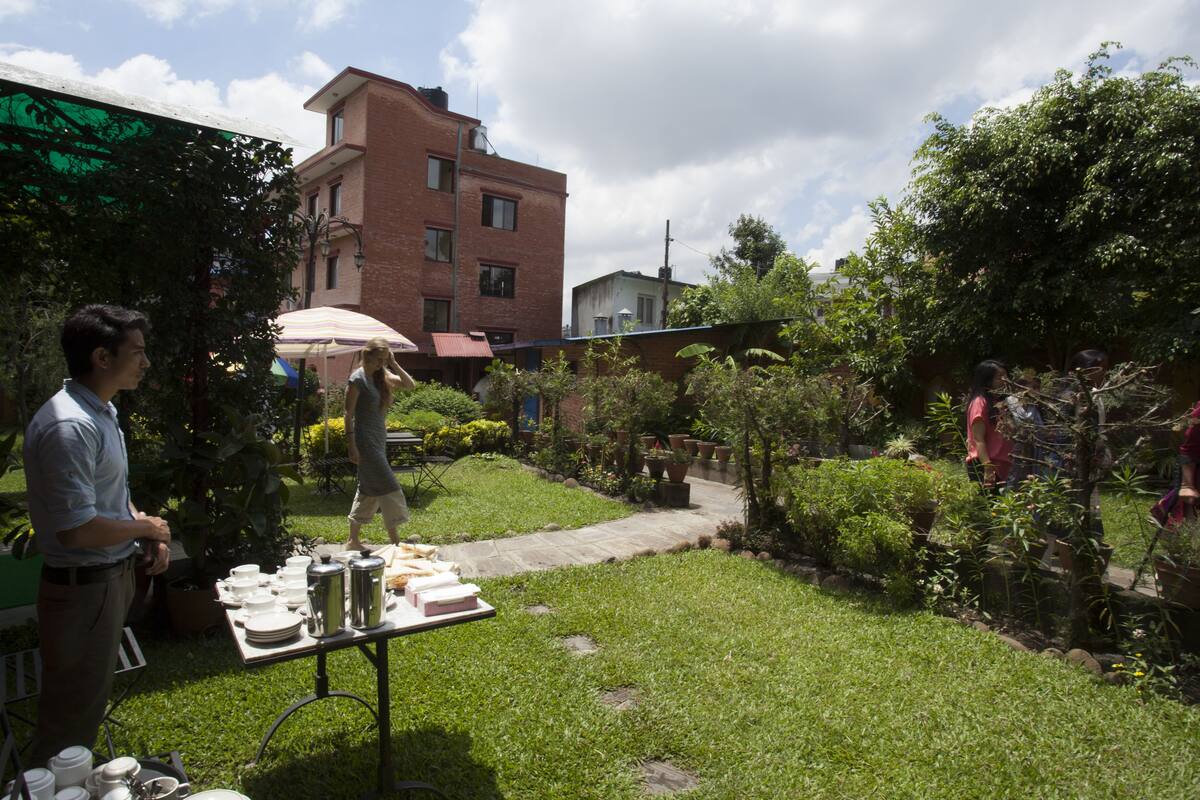 A garden with a table set up for a meal, surrounded by greenery and a brick building in the background.
