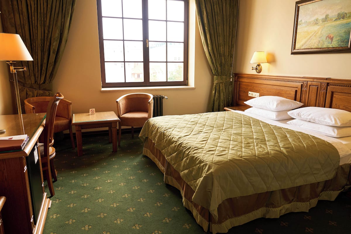 Habitación de hotel con cama, escritorio, silla, ventana con cortinas y un cuadro en la pared.