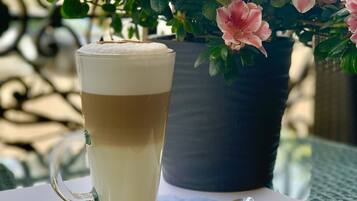 A glass of layered beverage on a saucer with a spoon, a menu, and a potted plant with pink flowers in the background.