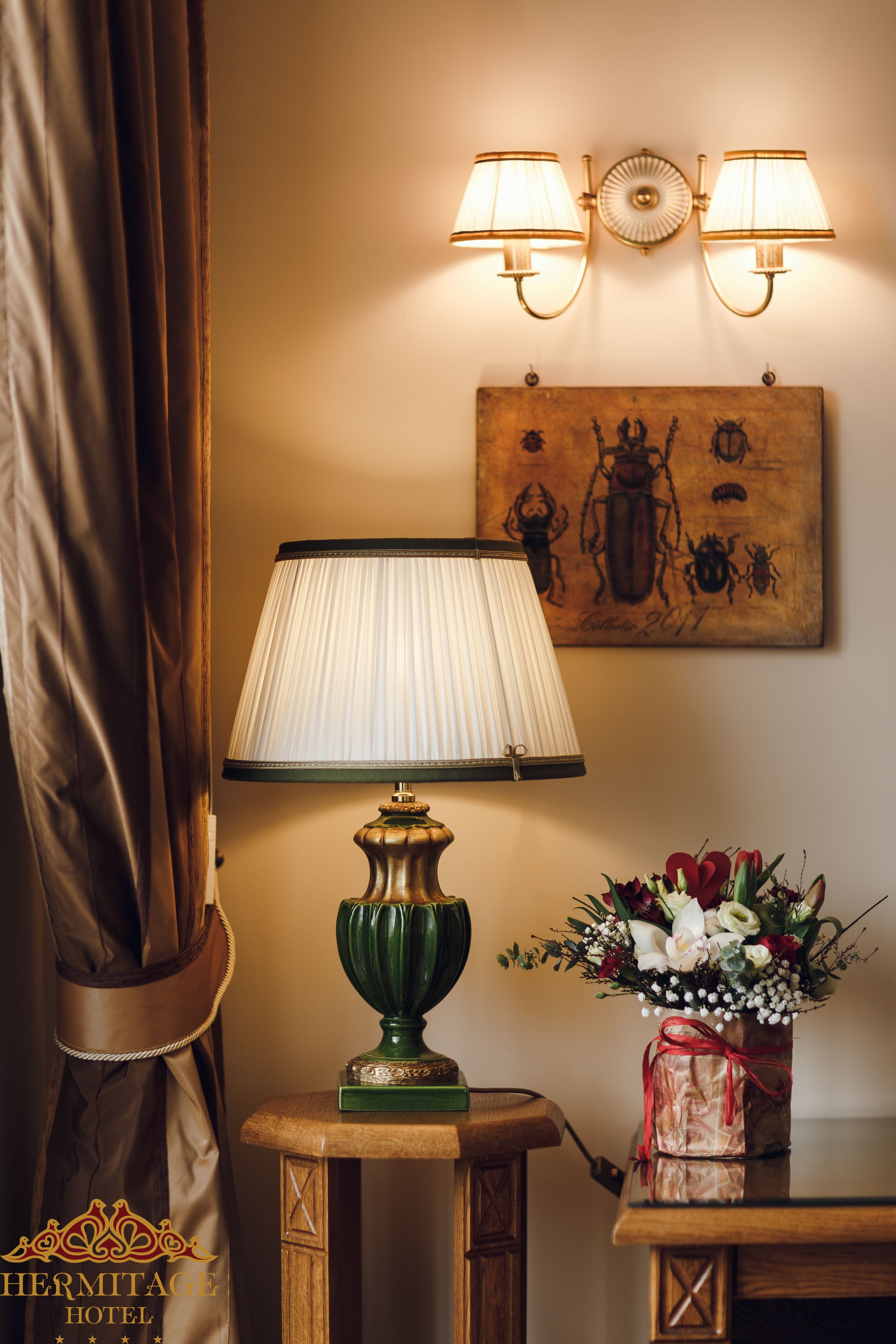 A lamp with a green base, a wall-mounted light fixture with two lamps, a painting of insects, and a vase with flowers on a wooden table.