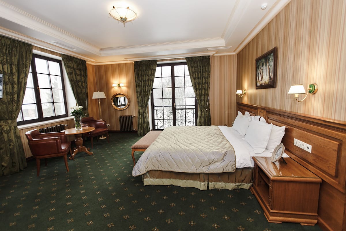 Habitación de hotel con una cama grande, cabecero de madera, mesita de noche, escritorio con silla, espejo redondo y ventana con cortinas.