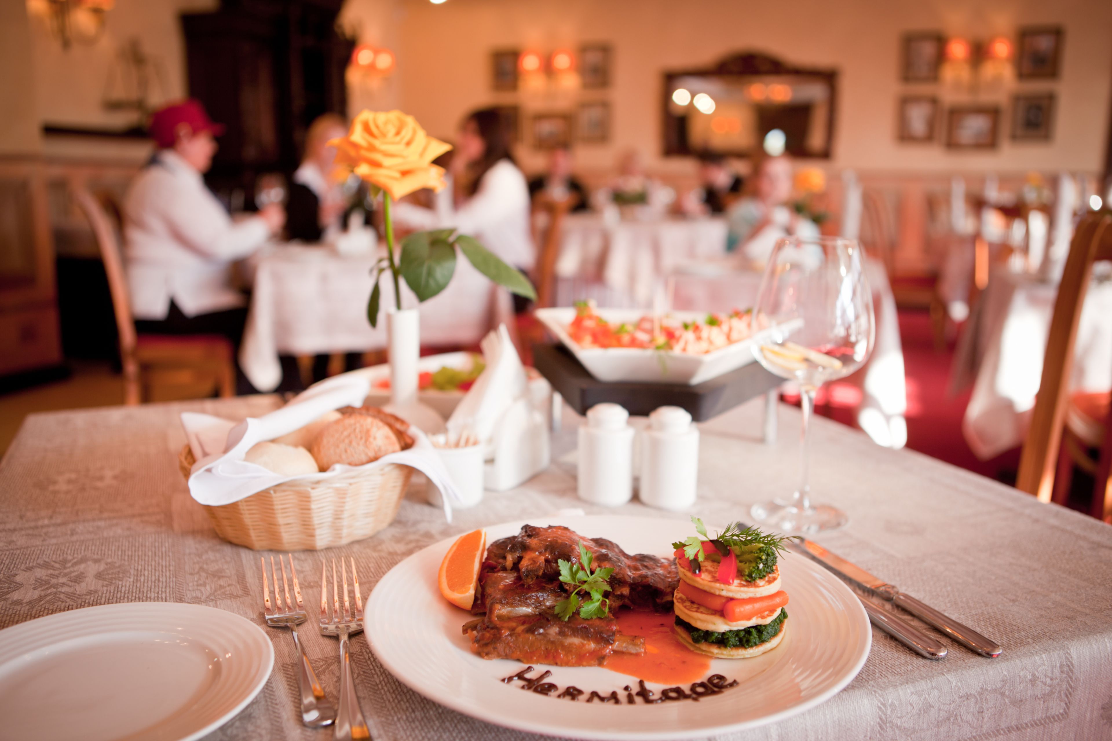 Una mesa de comedor dispuesta con un plato de comida, una rosa, saleros y pimenteros, una cesta de pan y una copa de vino.