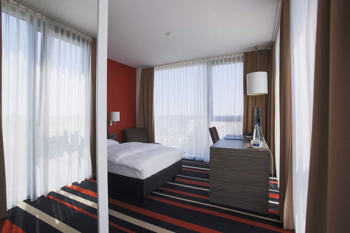 Quarto de hotel com cama, escrivaninha, cadeira, luminária e vista para a cidade.
