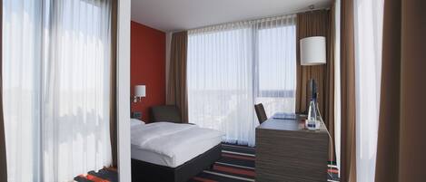 A hotel room with a bed, a desk, a chair, a lamp, and a view of the city.