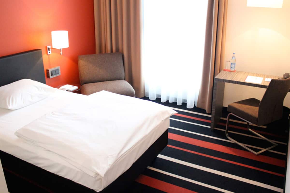 Quarto de hotel com cama, escrivaninha, cadeira, luminária e janela com cortinas.