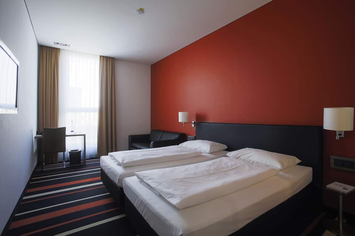 Quarto de hotel com duas camas, uma escrivaninha, uma cadeira e uma janela com cortinas.