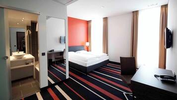 A hotel room with a bed, a desk with a phone, a TV, a bathroom with a sink, and a mirror.