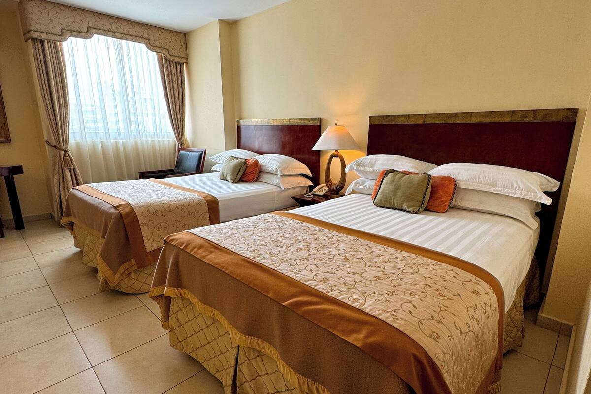 A hotel room with two beds, a nightstand, a lamp, and a window with curtains.