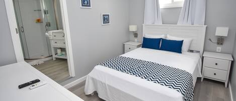 A neatly arranged bedroom with a bed, bedside tables, a nightstand, a desk, and a bathroom visible through an open door.