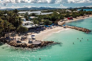 An aerial view of a beach resort with a rocky breakwater, clear turquoise waters, and a backdrop of mountains.