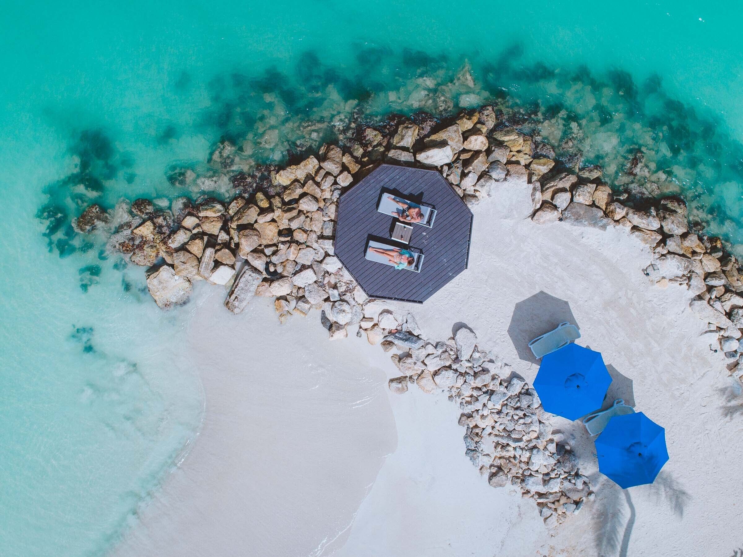 Vue aérienne d’une plage avec une terrasse en bois, deux parasols et un espace équipé de chaises longues.
