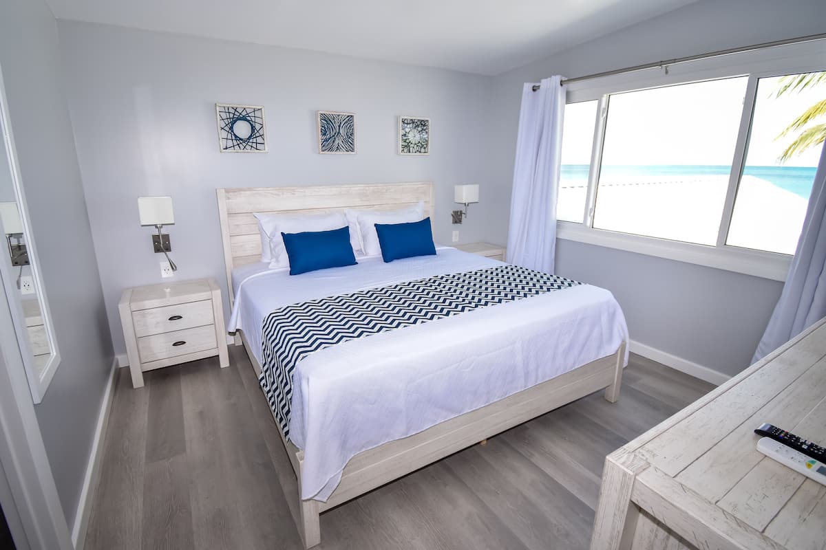 Une chambre à coucher comprenant un lit, des tables de chevet, une commode, une fenêtre donnant sur la plage et des œuvres d’art accrochées au mur.