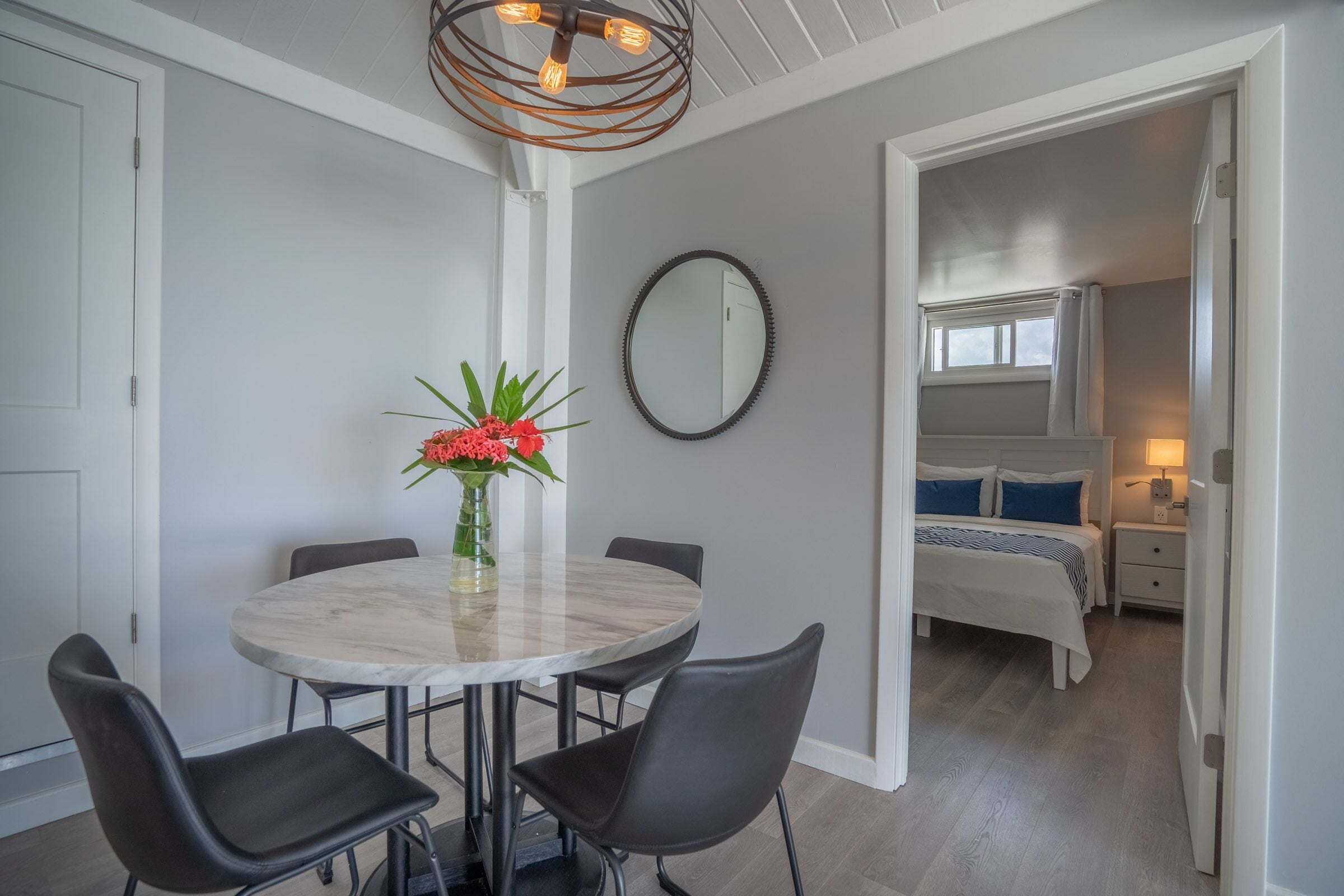 A room with a round marble table, two black chairs, a vase with red flowers, a round mirror, a bed with blue and white bedding, and a nightstand with a lamp.