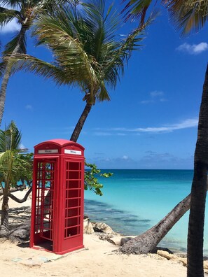 A red telephone booth on a beach with clear turquoise water and palm trees. - Siboney Beach Club (St. John's)