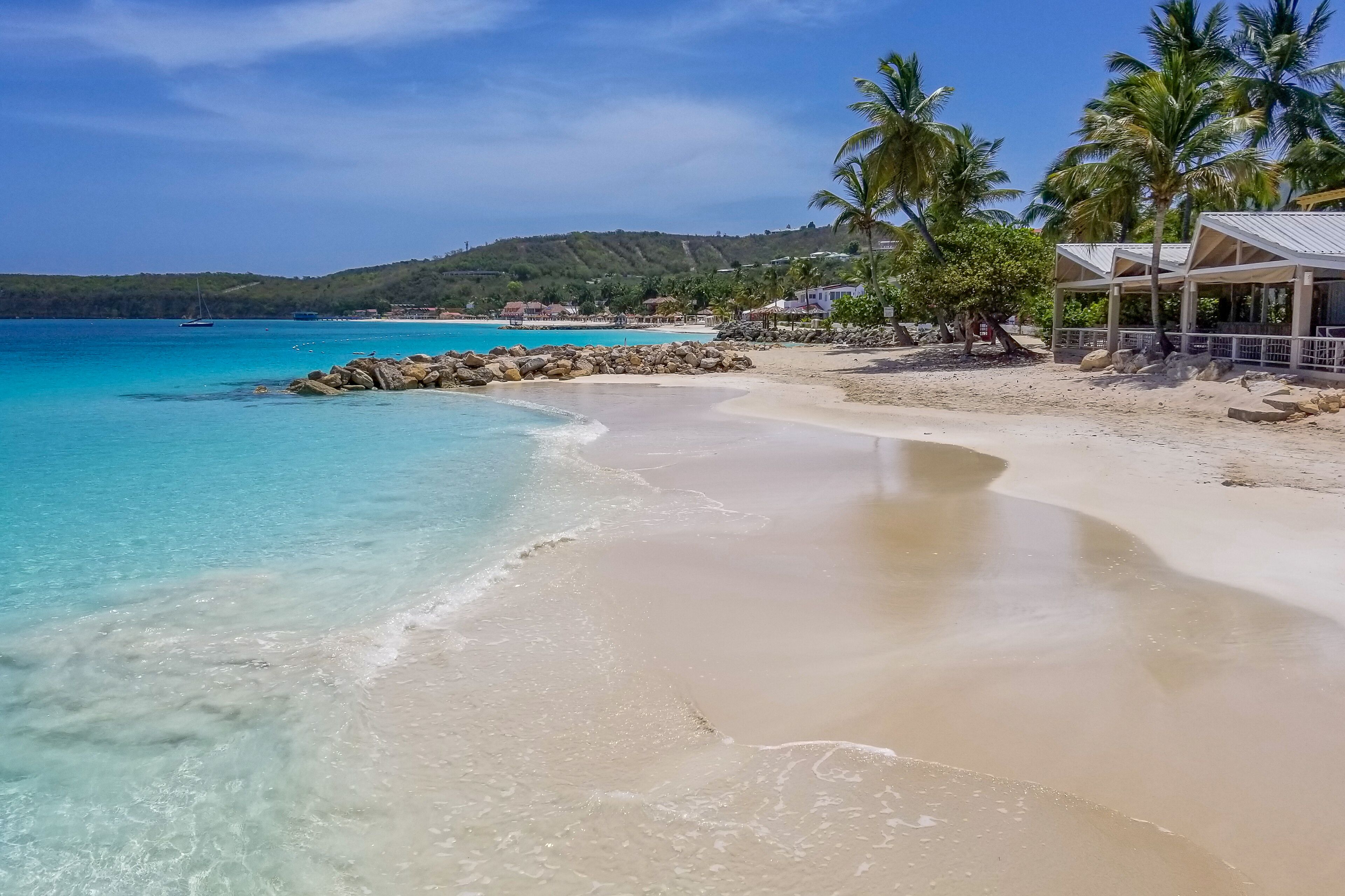 A beach with clear turquoise water, a sandy shore, and a structure with a roof and palm trees.