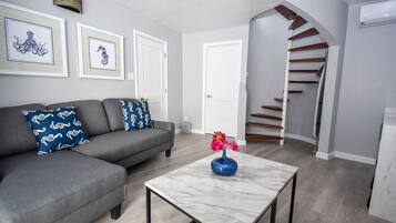 Una sala moderna con un sofá gris, una mesa de centro de mármol con un florero, y una escalera que conduce a un piso superior.