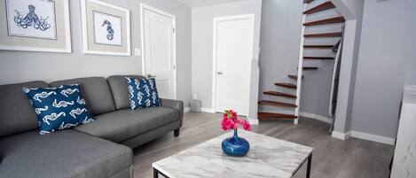 A modern living room with a grey sofa, a marble coffee table with a vase of flowers, and a staircase leading to an upper floor.