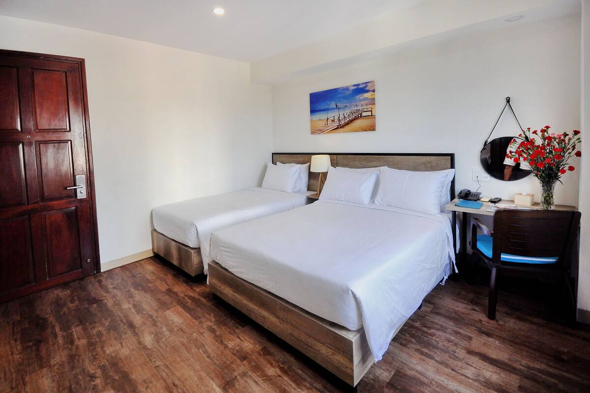 A hotel room with two beds, a wooden floor, a desk with a chair, a painting on the wall, and a door.