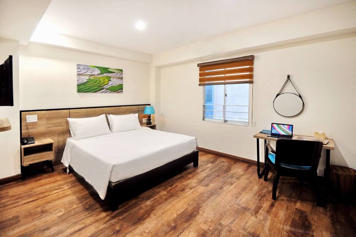 A hotel room with a wooden floor, a bed with white linens, a desk with a laptop, a chair, a window with blinds, and a wall-mounted mirror.