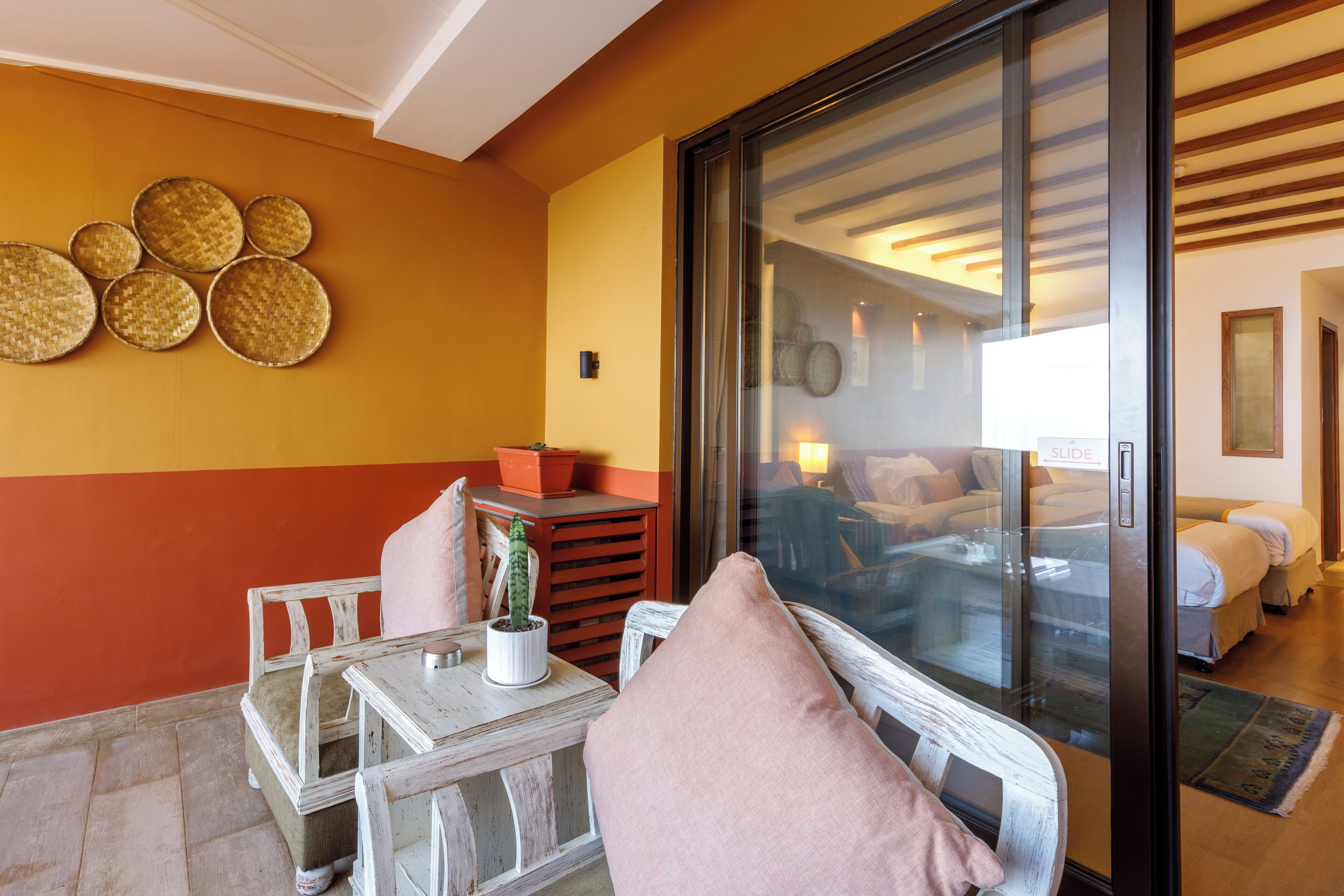 A room with a large sliding glass door leading to a bedroom, a small table with a potted plant, and a wall with circular woven decorations.