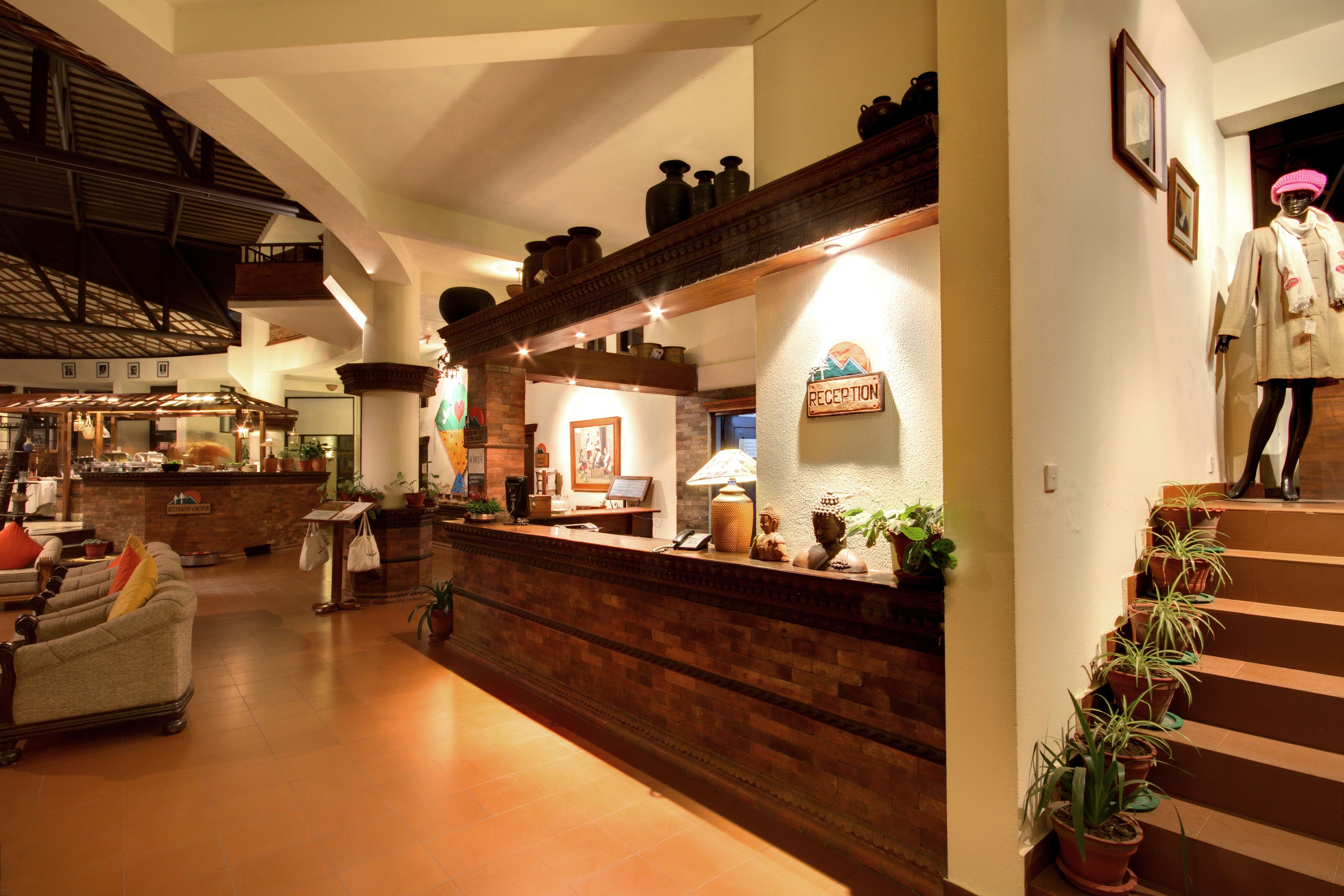 Un área de recepción con un mostrador de madera, un maniquí vestido con abrigo y sombrero, y una escalera decorada con plantas en macetas.