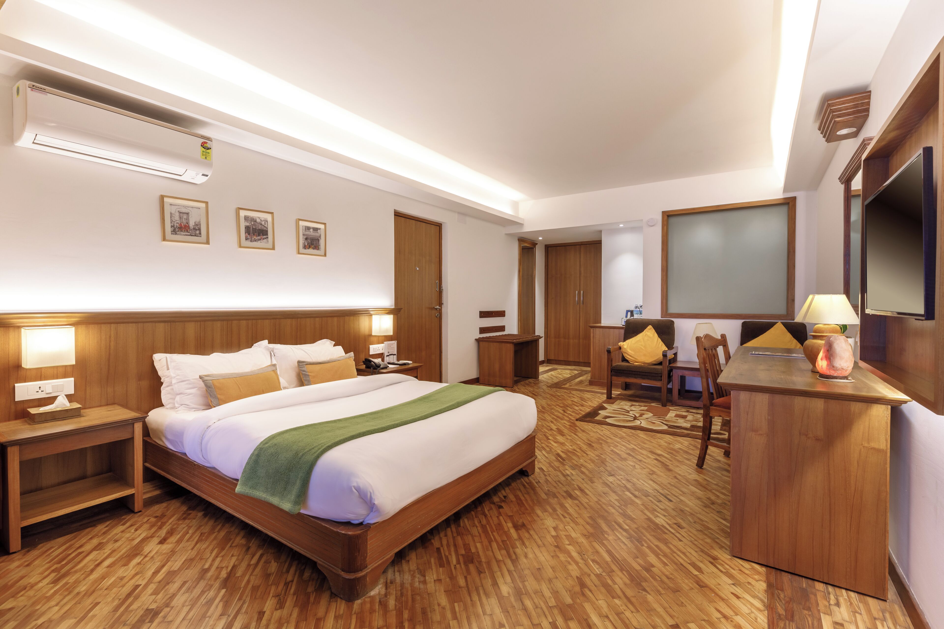 A hotel room with a wooden bed, bedside tables, a desk with a chair, a TV, and framed pictures on the wall.