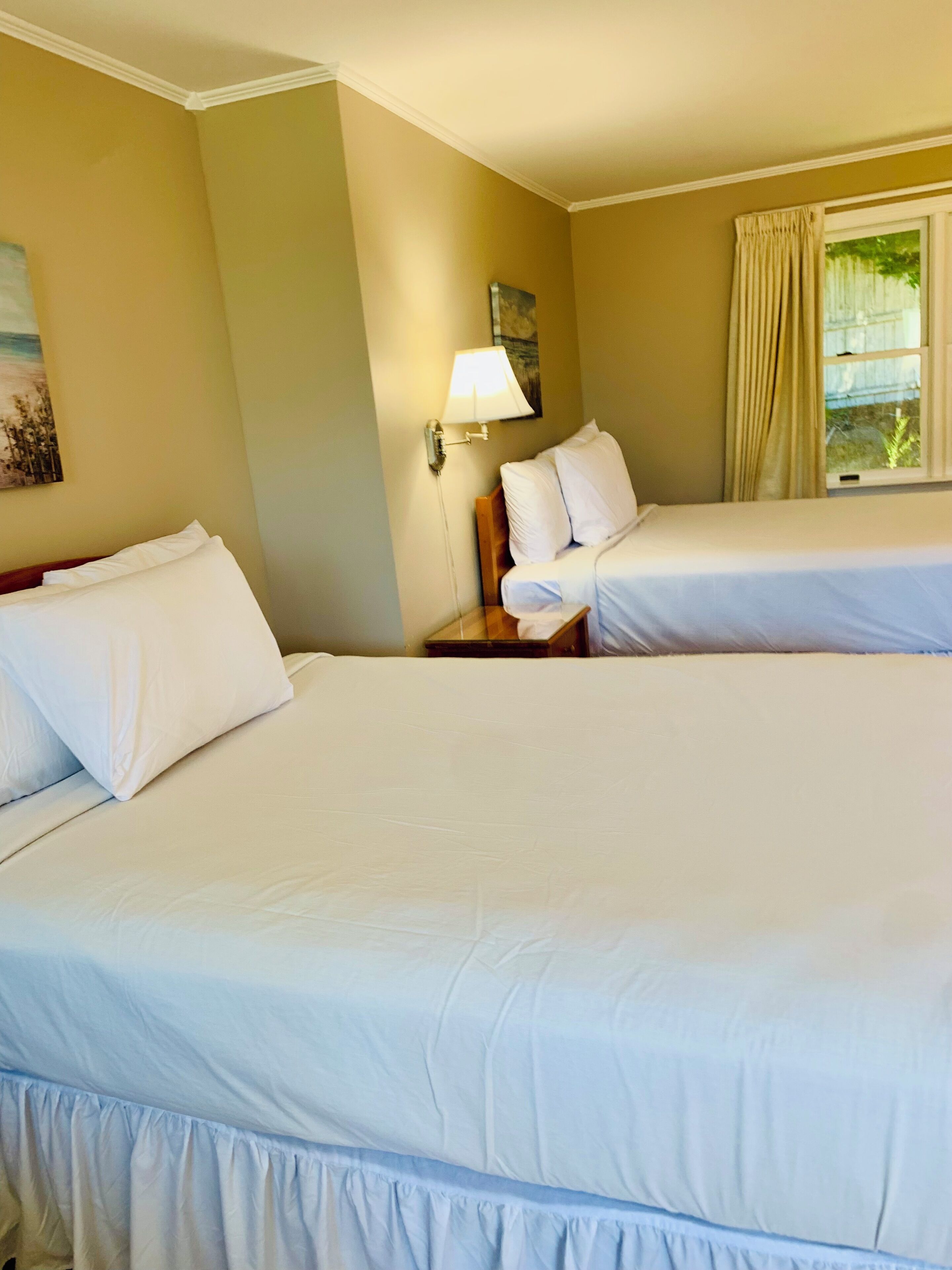 A hotel room with two beds, a nightstand, a lamp, and a window with curtains.